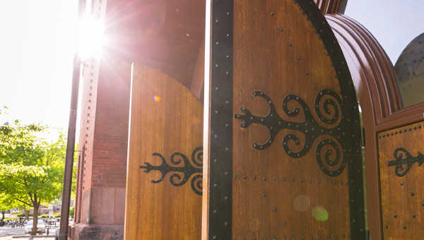 Close-up detail image of the arched wooden doors of the Red Gym, 
                        with ornate decorative hinges, swung open with the sun shining in the background